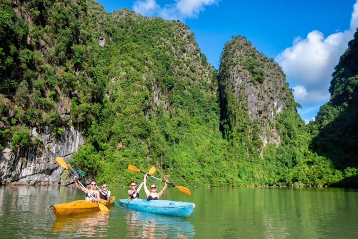 Travellers paddling yellow and blue kayaks past jungle-covered limestone cliffs on calm green water in Vietnam