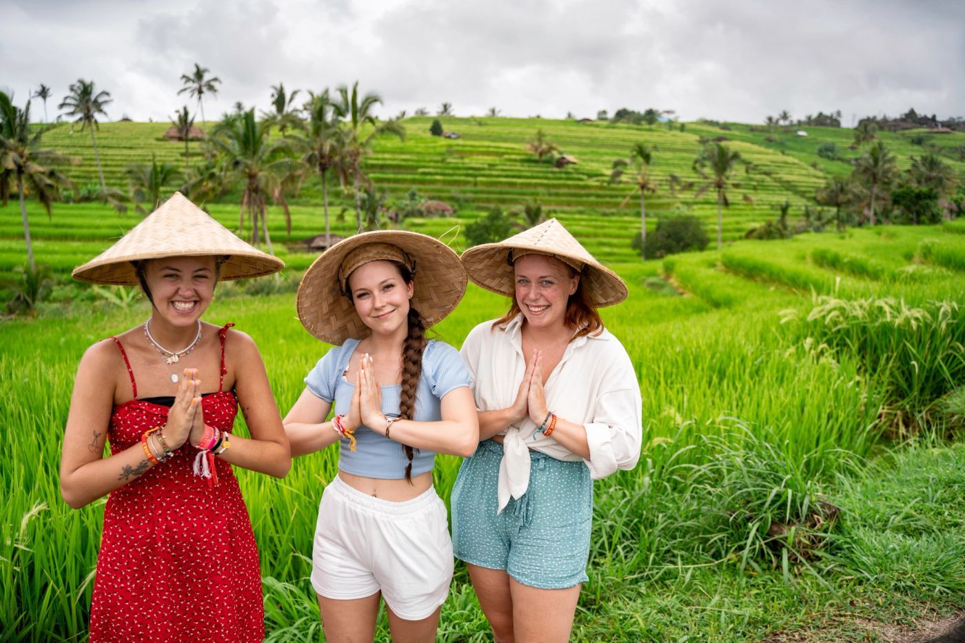 Three travellers in conical hats smiling with hands pressed together in front of green Bali rice terraces
