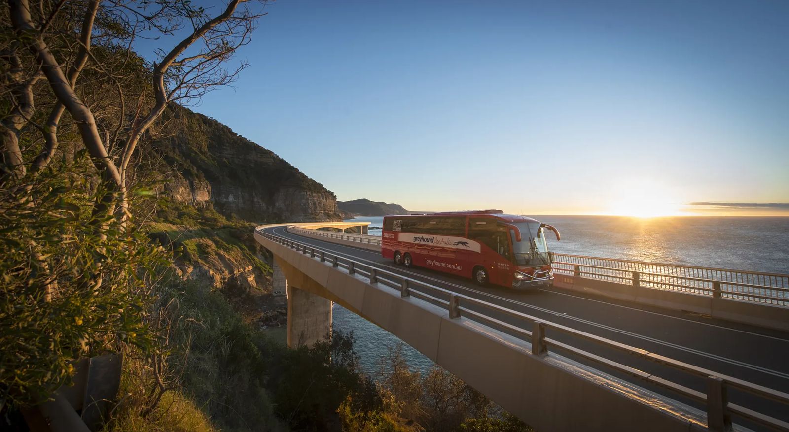 A red Greyhound coach crossing a curved coastal bridge beside sandstone cliffs, with the sun rising over the ocean
