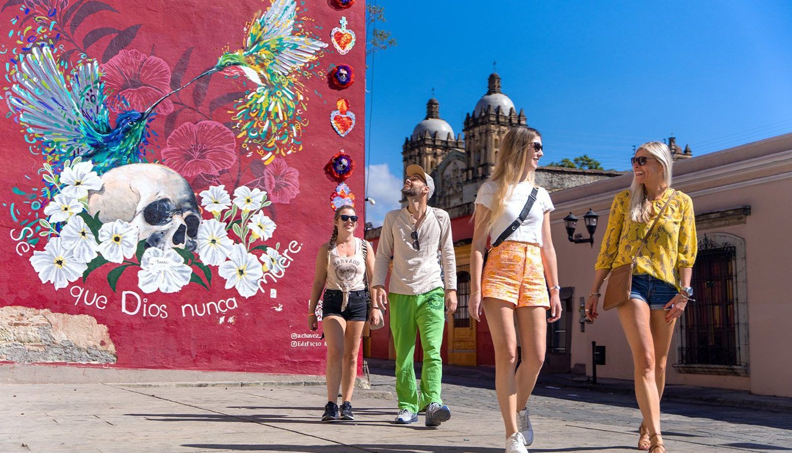 Four young travellers walking past a bright red hummingbird and skull mural on a cobbled street in Oaxaca, Mexico, with a baroque church behind them