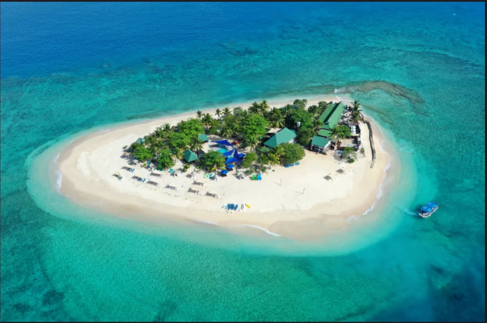 Aerial view of a small Fijian island ringed by white sand and turquoise reef, with palm trees, bures and a boat pulling up to the beach