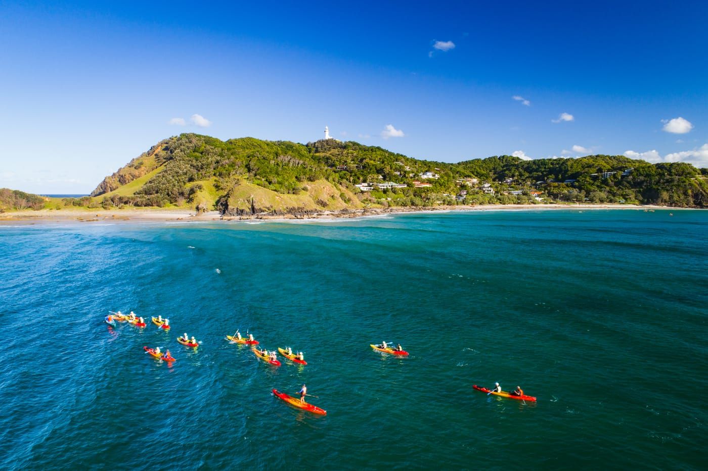 Aerial view of a group of sea kayaks paddling on deep blue water off Byron Bay, with a green headland and the Cape Byron lighthouse behind