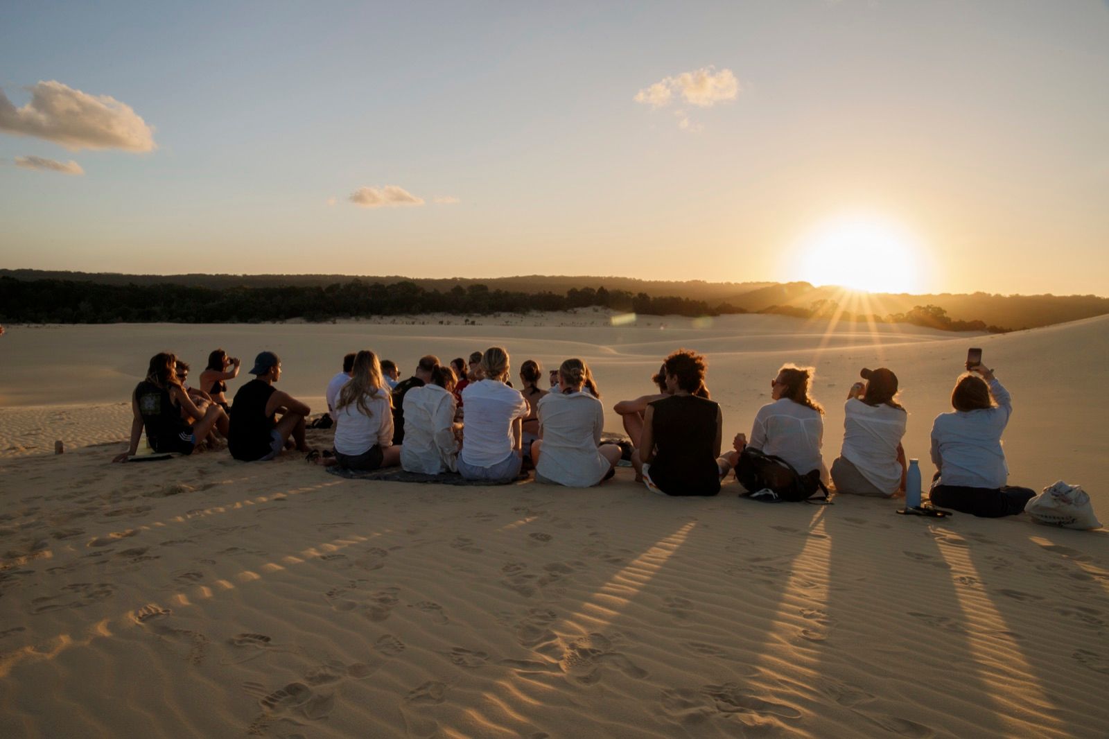 A group of travellers sitting on a sand dune on K'gari at sunset, with the sun low over the treeline and long shadows across the sand