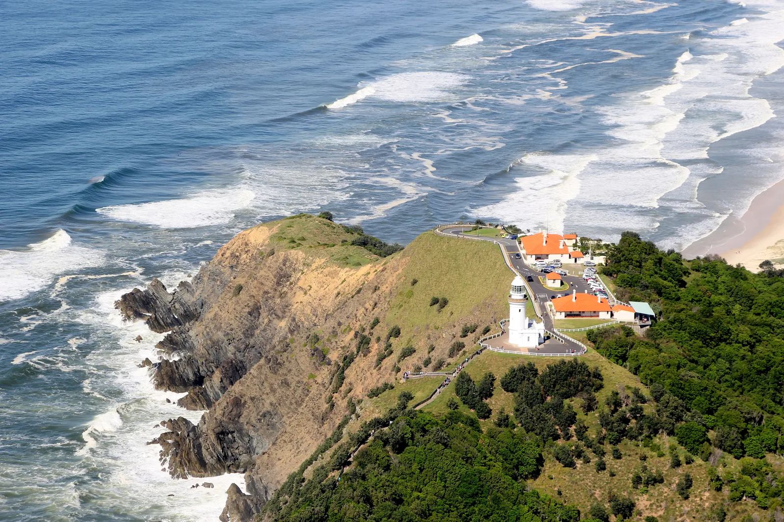 Aerial view of the white Cape Byron lighthouse on a green headland, with lines of surf breaking on the beaches either side
