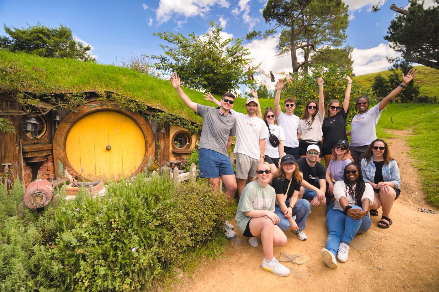 A group of young travellers with arms raised outside a round yellow hobbit-hole door on the Hobbiton movie set