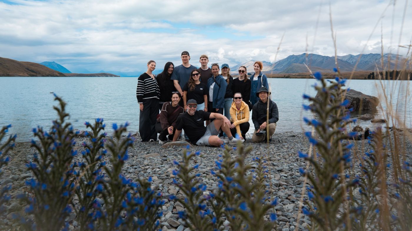 A group of young travellers posing together on a pebbly lake shore with blue flowers in the foreground and mountains behind the water