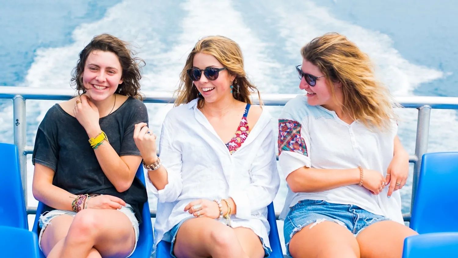 Three travellers laughing on the open top deck of the island catamaran, with its white wake stretching out behind them