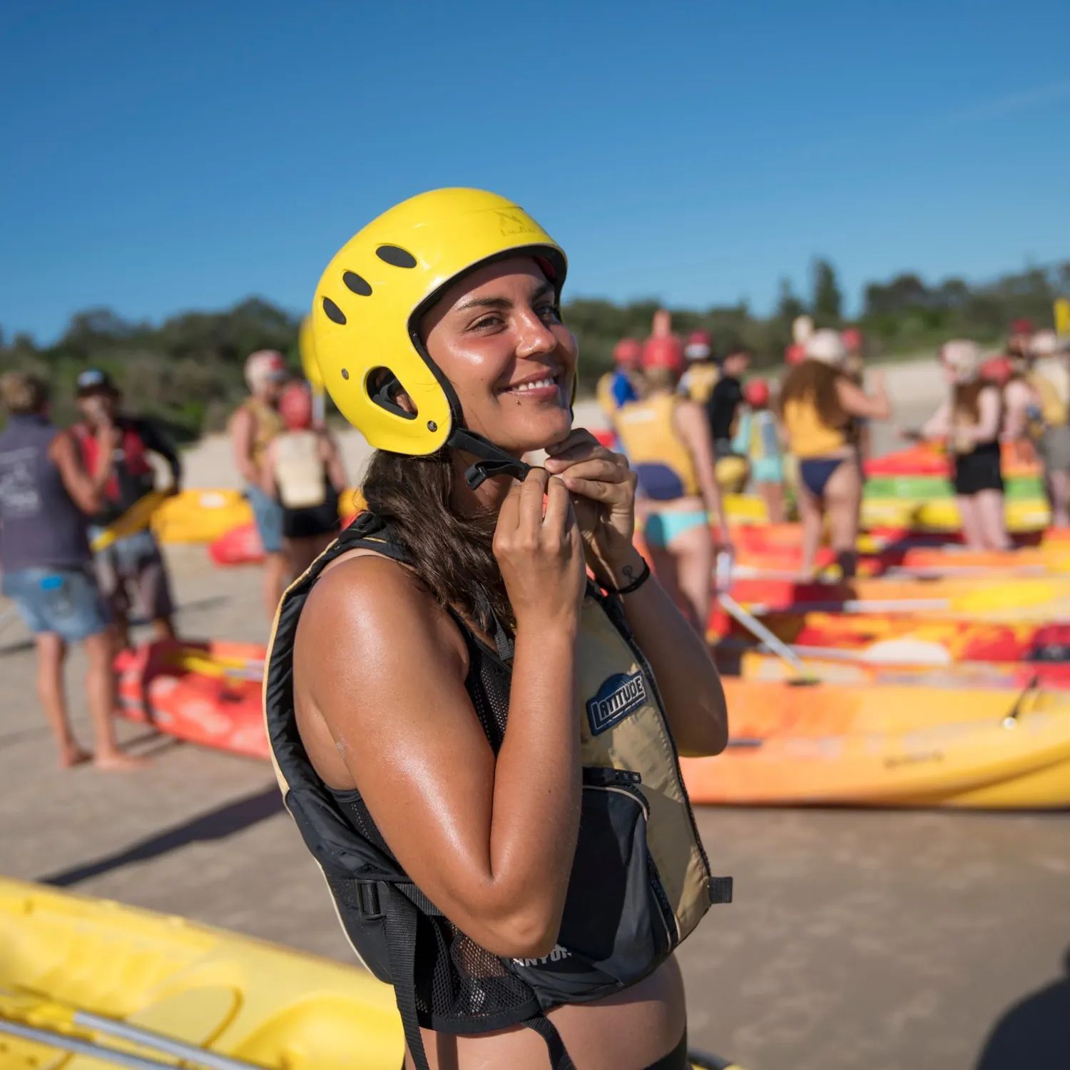 Smiling paddler in a yellow helmet and life vest on a beach, with kayaks and a group behind her