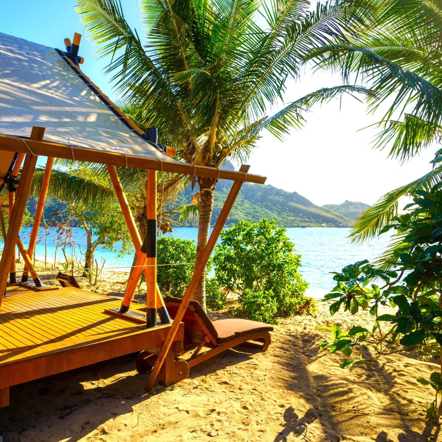 A shaded beach daybed under palm trees on Kuata, looking across turquoise water to the green peaks of the Yasawa Islands