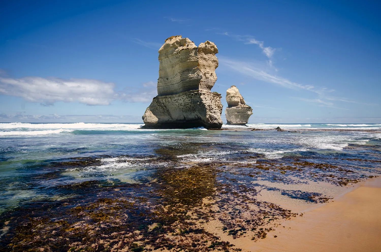 Limestone rock stacks of the Twelve Apostles standing in the surf beside a sandy beach under a blue sky