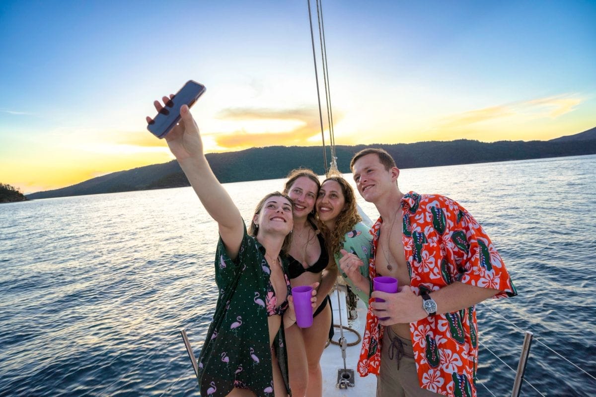 Four young travellers taking a selfie on the deck of a sailing boat at sunset, with forested islands behind them
