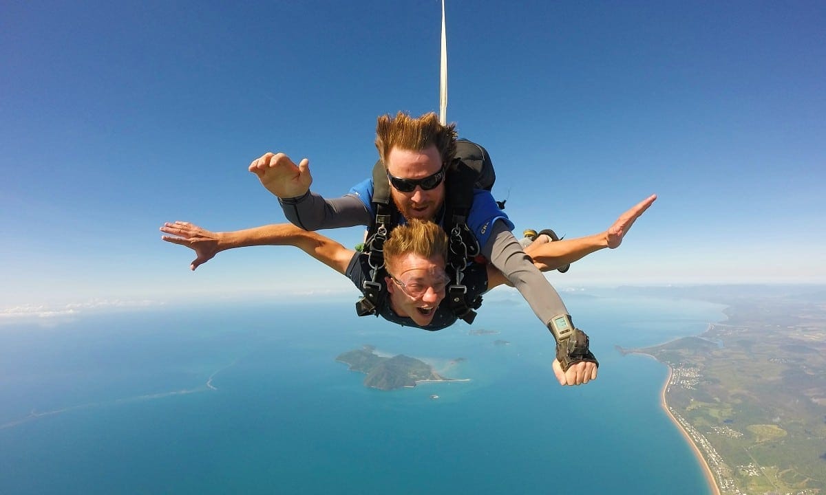 A tandem skydiver and instructor in freefall with arms spread, above a blue coastline with a small island and a long beach below