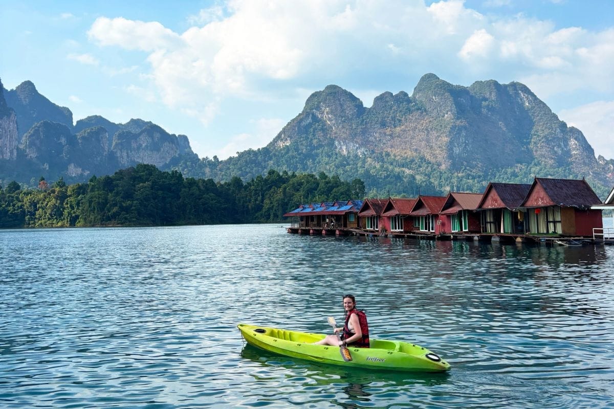 A traveller paddling a green kayak on a lake, with a row of red floating bungalows beside tree-covered limestone mountains