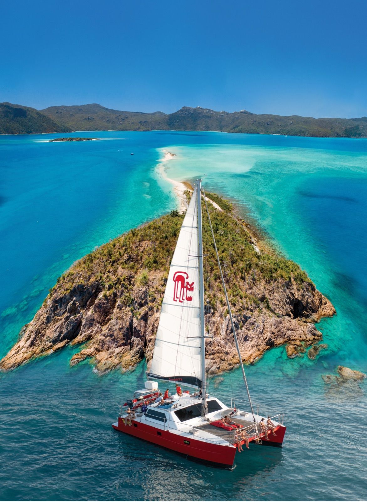 Aerial view of a sailing catamaran anchored beside a small rocky island with a white sand spit, surrounded by turquoise Whitsundays water