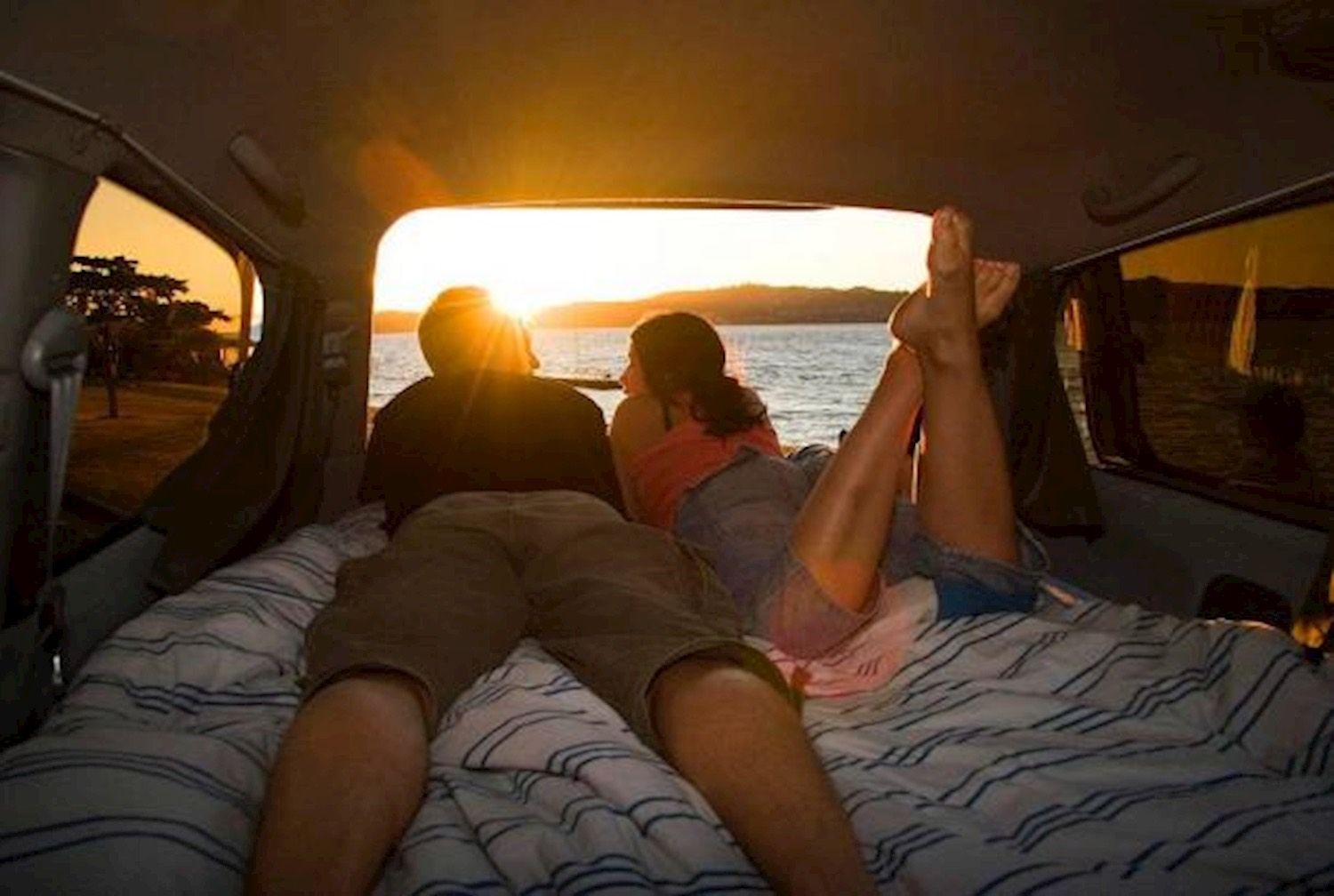 Two travellers lying in the back of a campervan watching the sun set over the water