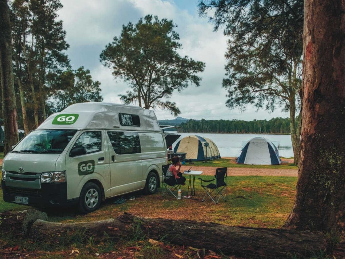 A white hi-top campervan parked at a lakeside campground beside two tents